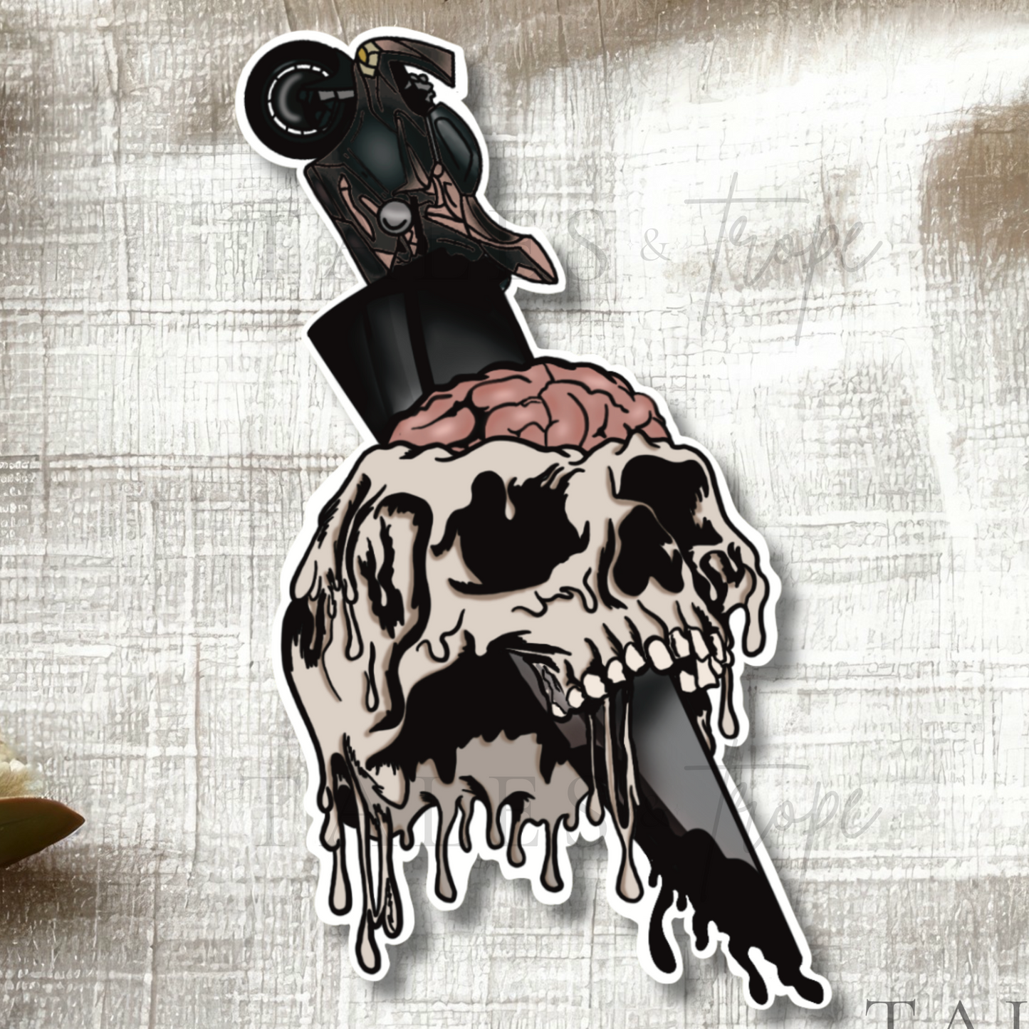 A die-cut sticker featuring a dripping skull with a pink brain and a motorcycle on a knife, styled on a textured beige fabric with eucalyptus leaves. The brand name 'TALES & Trope' is visible in the bottom right corner.