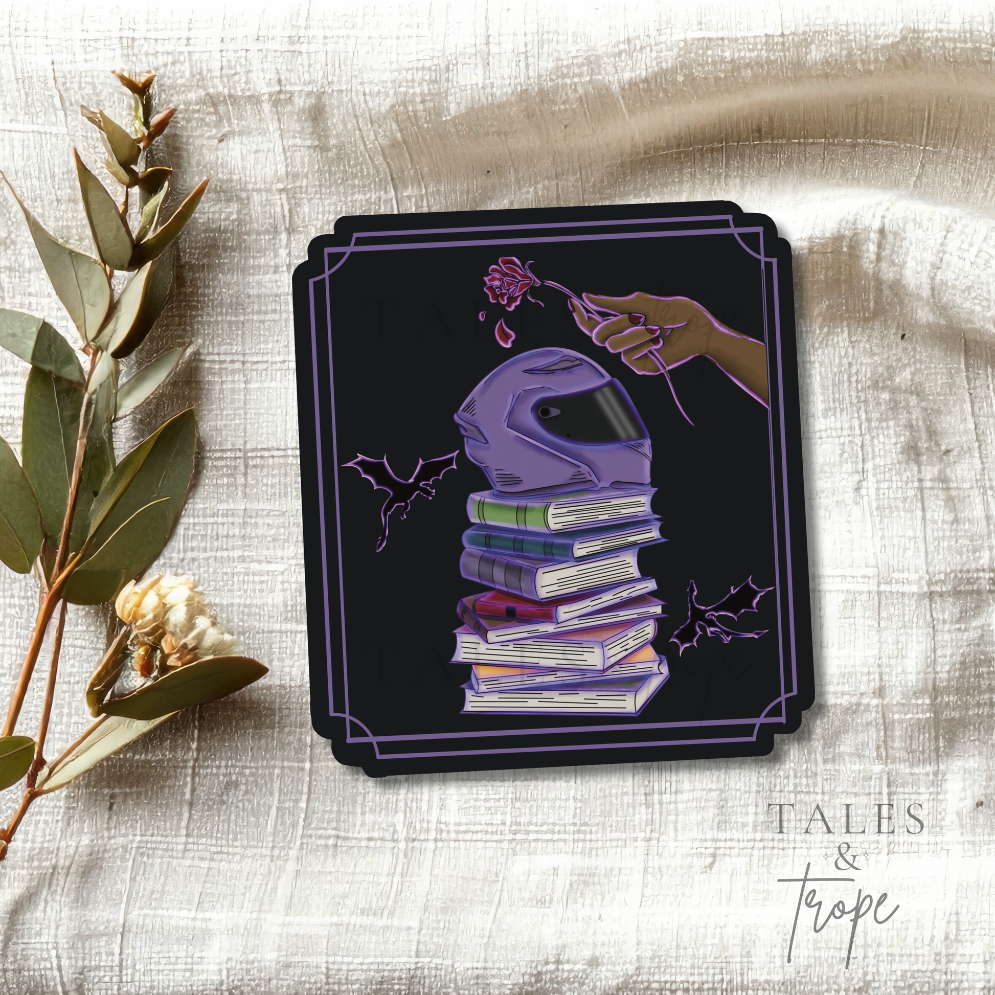 A square black art print or coaster featuring a purple motorcycle helmet stacked on books with a hand holding a flower, styled on a textured beige fabric with dried eucalyptus branches. The brand name 'Tales & Trope' is visible in the bottom right corner.