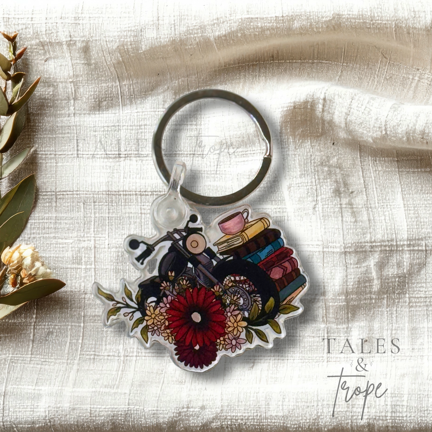 A die-cut acrylic keychain featuring a vintage motorcycle, books, coffee cups, and flowers, displayed on a textured cream linen fabric with flowers on the side.