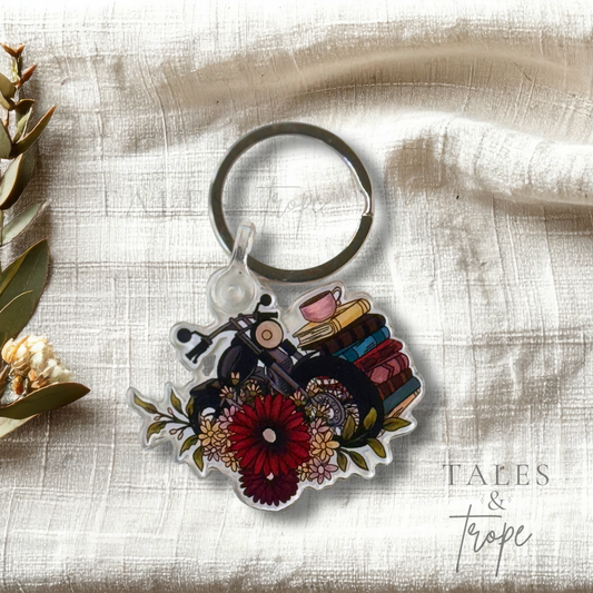A die-cut acrylic keychain featuring a vintage motorcycle, books, coffee cups, and flowers, displayed on a textured cream linen fabric with flowers on the side.