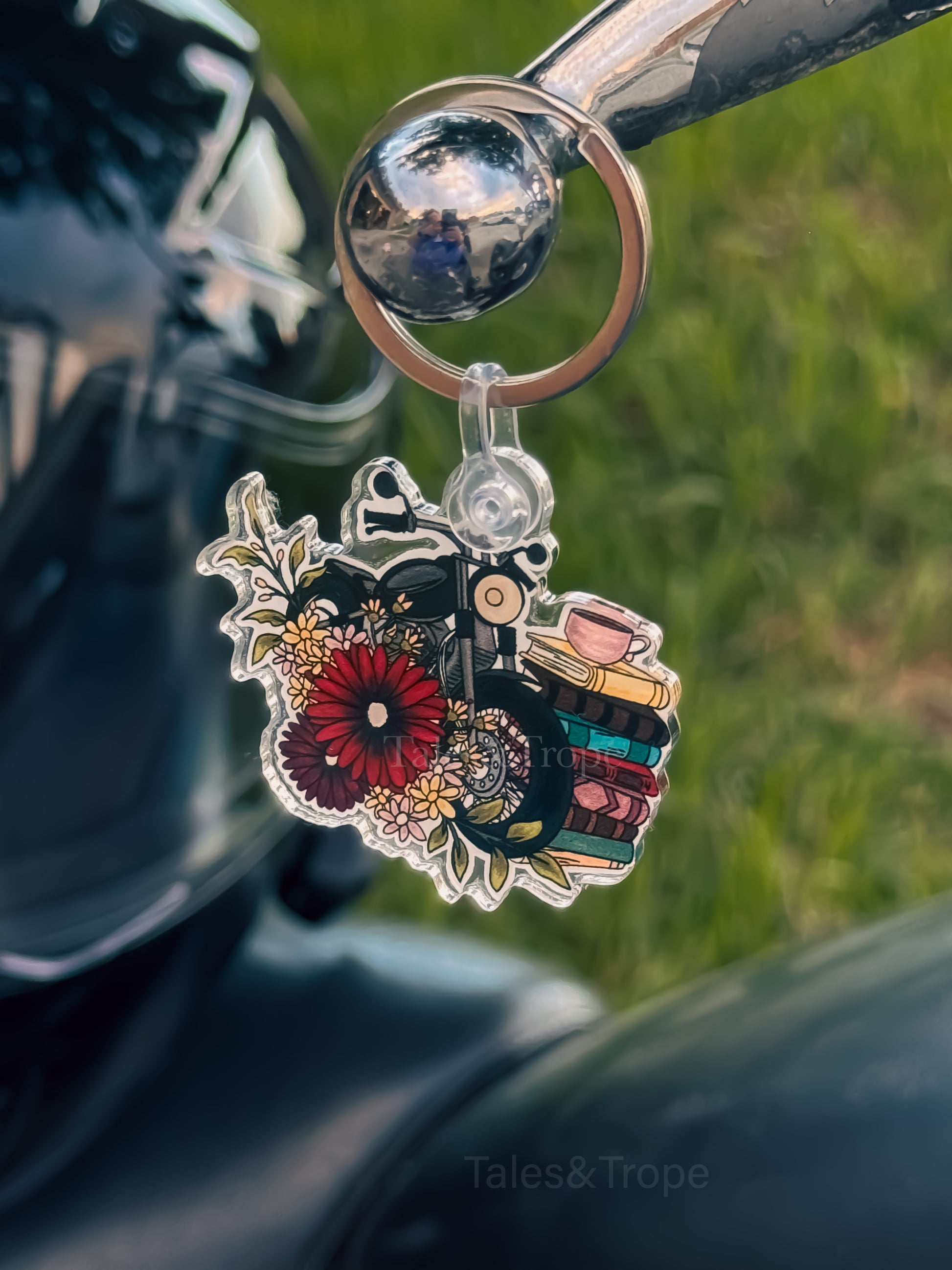 A close-up of a decorative keychain featuring a vintage bicycle, flowers, and books, hanging from a motorcycle handlebar with a blurred outdoor background.