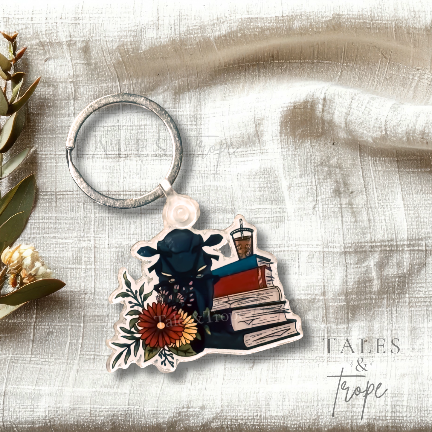 A die-cut acrylic keychain featuring an illustration of a black sportsbike on top of stacked books next to a red flower, attached to a silver metal key ring and displayed on a textured white fabric background.
