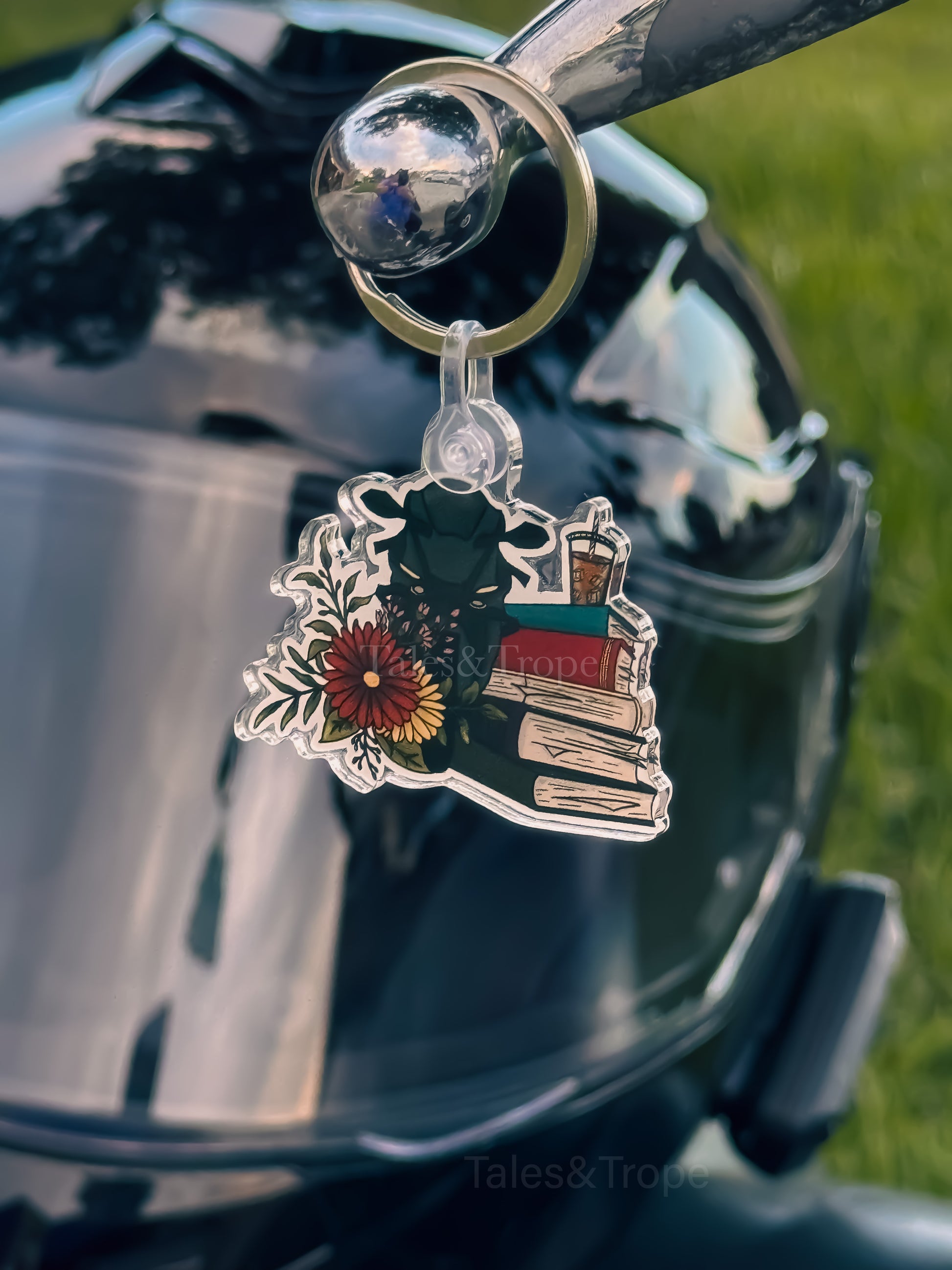 A clear acrylic keychain charm of a sports bike, books, and a flower, hanging from a metal ring on a motorcycle helmet.