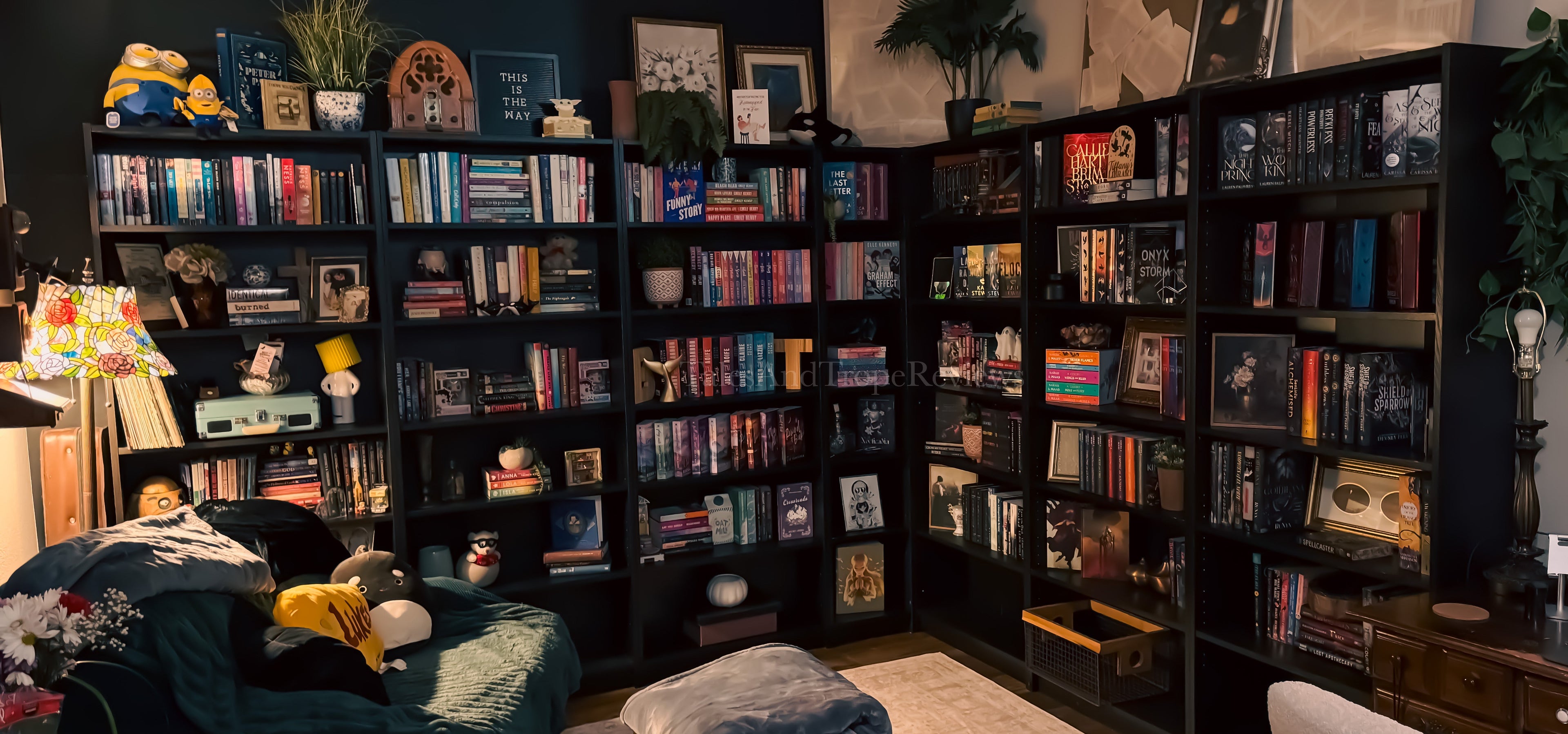 A cozy home office or living room corner featuring large black corner bookshelves filled with books, framed art, plants, and decorative items. A dark green recliner sits on the left, a wooden chest with a folded blanket in the center, and a desk with a computer monitor on the right.
