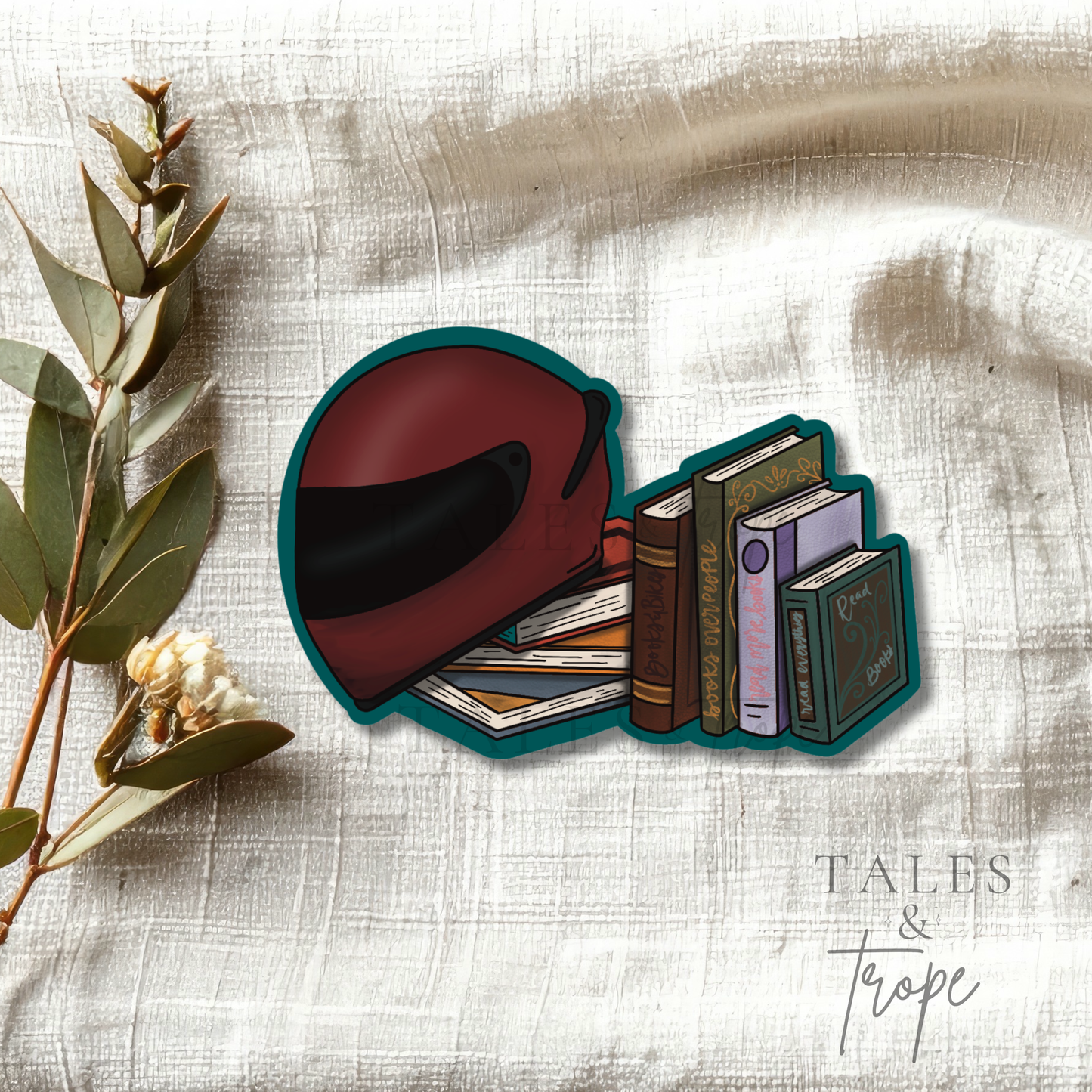 A die-cut sticker featuring an illustration of a dark red motorcycle helmet and a stack of books, placed on a textured beige fabric background with dried eucalyptus leaves. The brand name 'TALES & Trope' is visible in the bottom right corner.