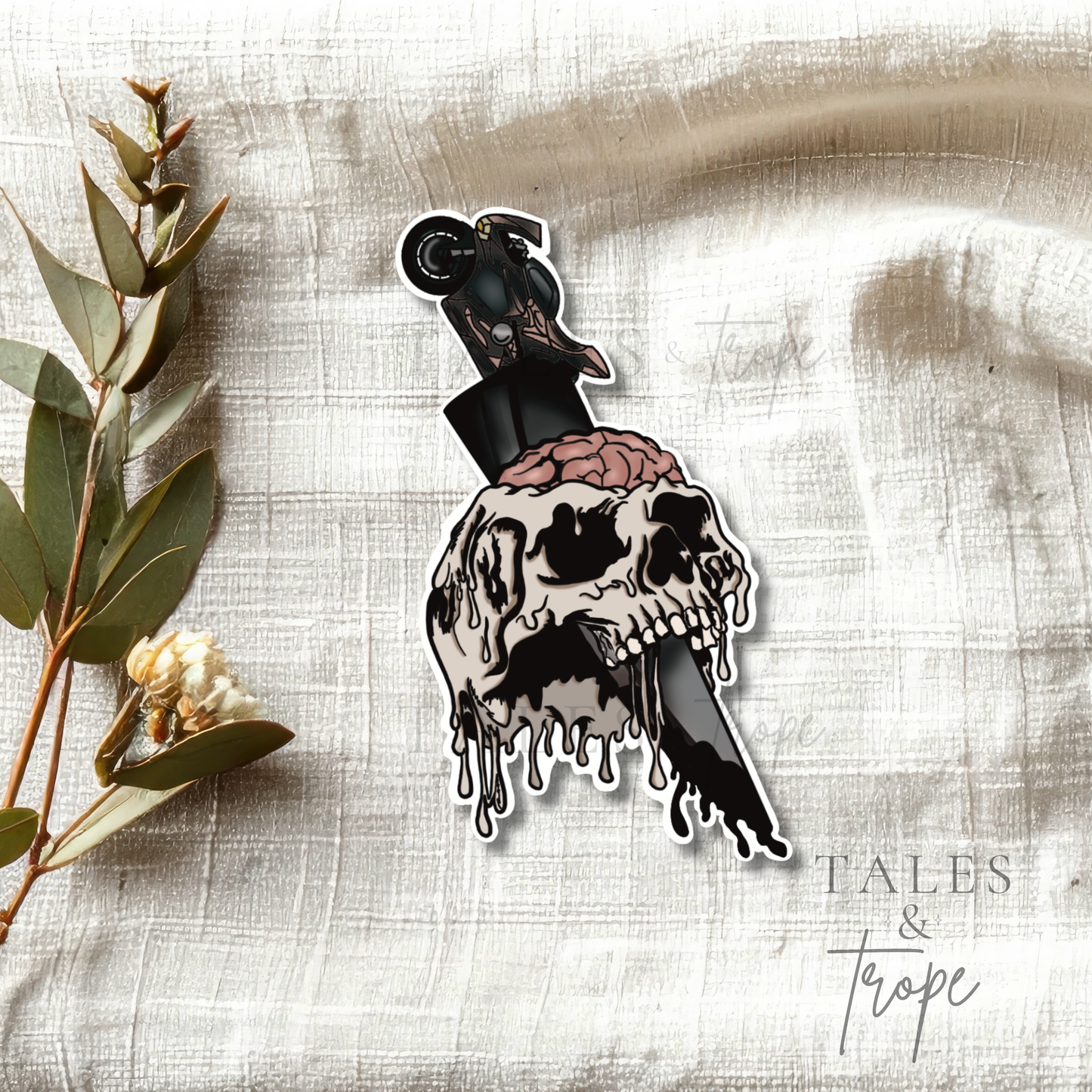A die-cut sticker featuring a dripping skull with a pink brain and a motorcycle on a knife, styled on a textured beige fabric with eucalyptus leaves. The brand name 'TALES & Trope' is visible in the bottom right corner.