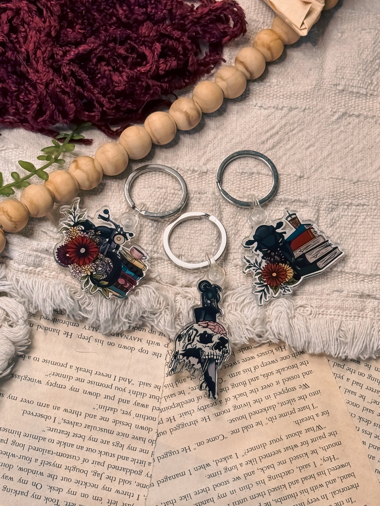 A styled flat-lay of four colorful acrylic keychains with motorcycle, floral and book illustrations, arranged on a textured cream fabric with a wooden bead garland, a burgundy knit piece, and an open book.